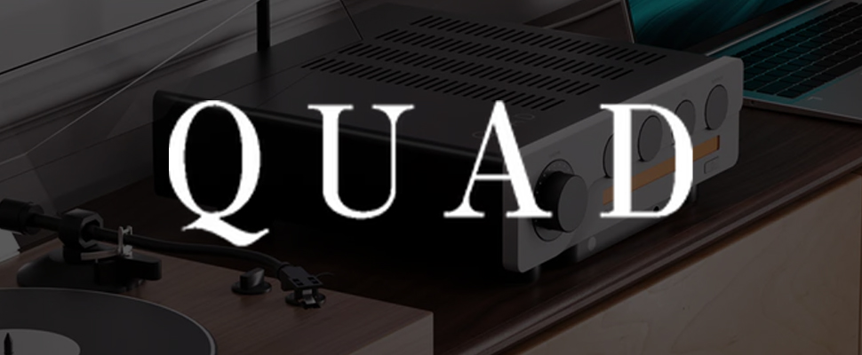 A stereo amplifier with dials and vents sits on a wooden table near a record player and an open laptop. The word "QUAD" is prominently displayed.