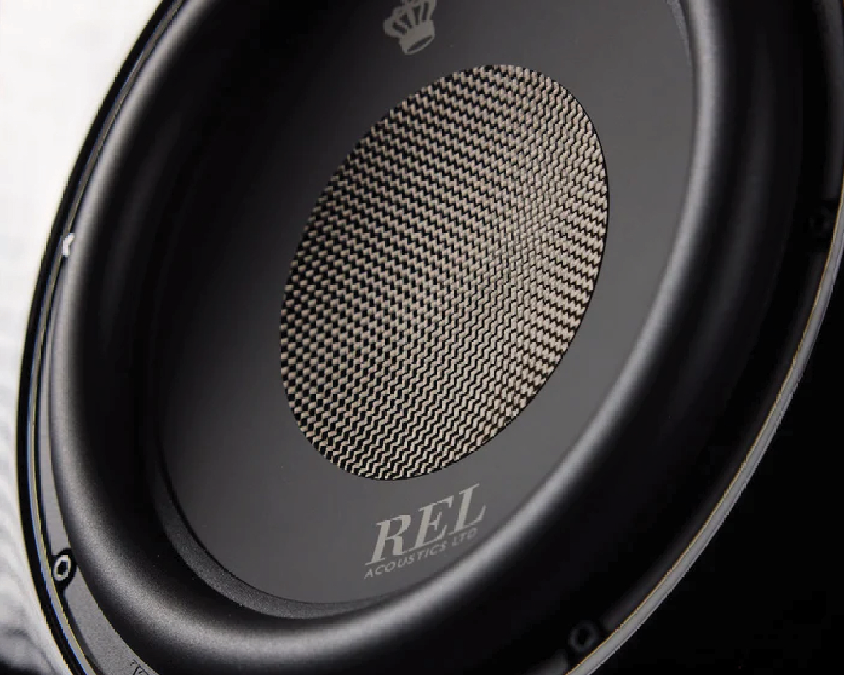 Close-up of a REL subwoofer speaker. The circular speaker features a woven pattern on the cone, bordered by a sleek black frame, conveying modernity.