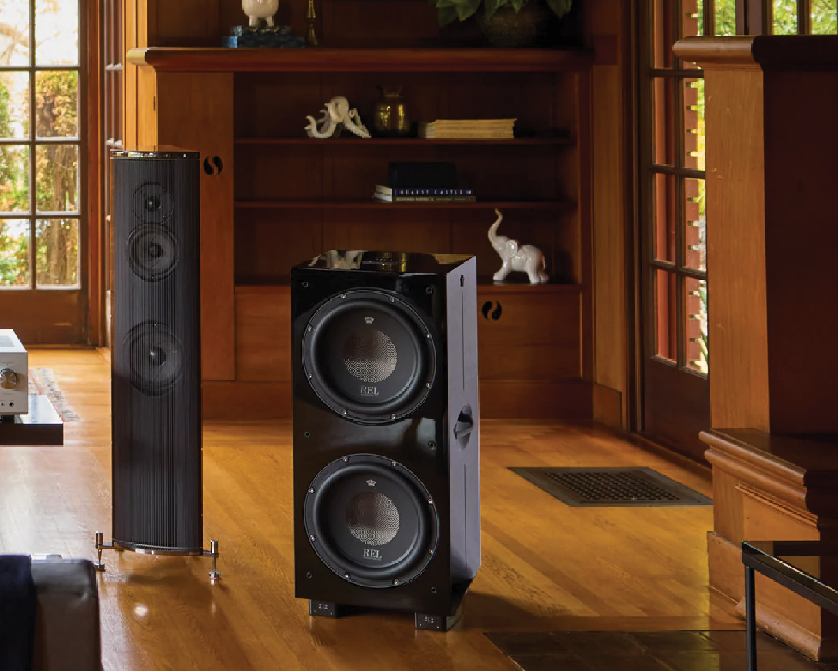 Modern living room with two large speakers, one tall and slim, one cubical with dual subwoofers. Warm wooden tones and elegant decor create a cozy, refined atmosphere.