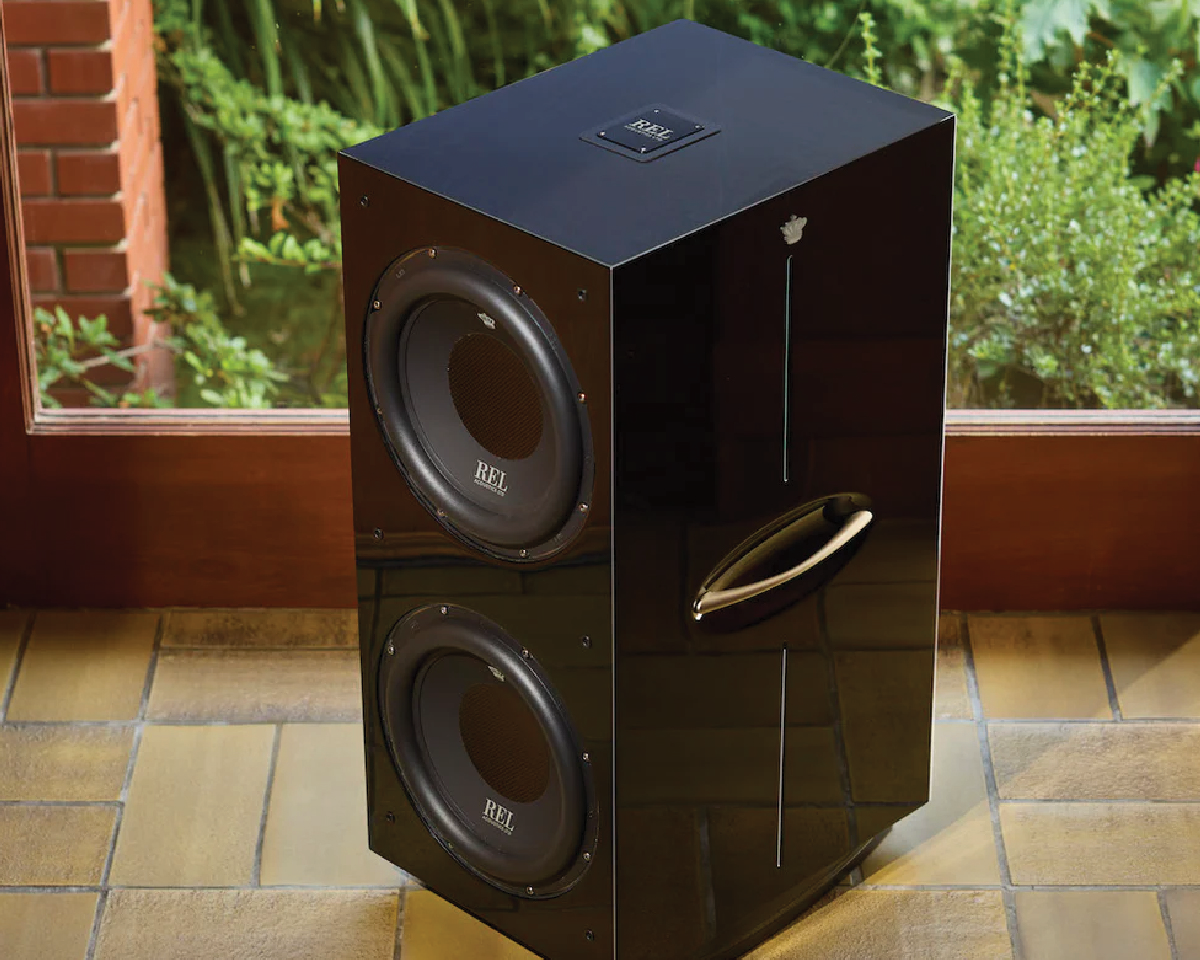 A sleek, black subwoofer with dual speakers sits on a tiled floor near a glass window. The room has a warm, natural ambiance with greenery visible outside.
