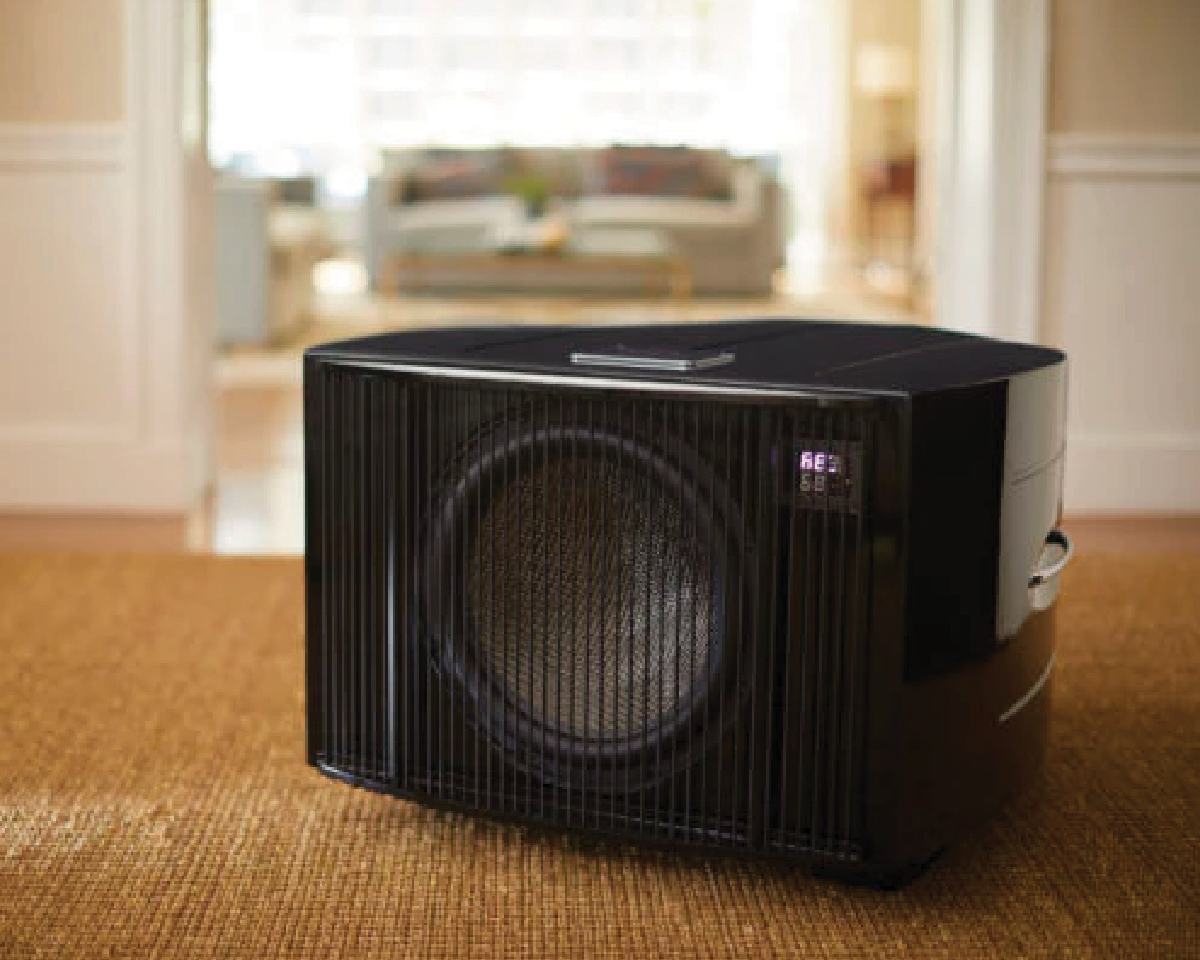 A sleek black portable air conditioner sits on a beige carpet in a bright, spacious room. Its digital display shows settings. The mood is modern and comfortable.
