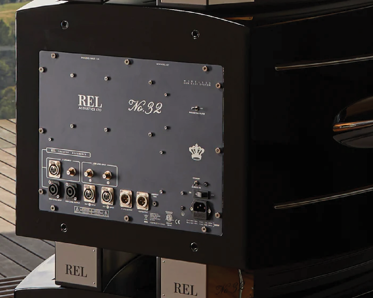 Close-up of a black REL Acoustics subwoofer with model "No. 32" on its back panel. Visible are various input/output ports and labels, set in an elegant room.
