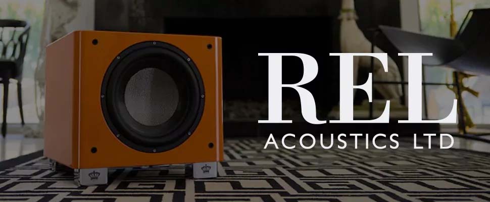 Orange subwoofer on a geometric-patterned rug in a modern living room, featuring large "REL ACOUSTICS LTD" text. The atmosphere is stylish and sophisticated.