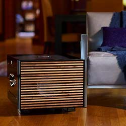A modern wooden cube-shaped space heater with horizontal slats sits on a hardwood floor beside a gray sofa with a blue throw, creating a cozy atmosphere.
