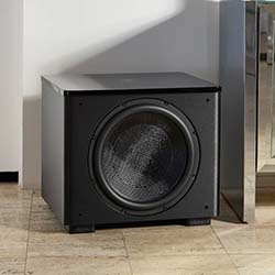 A black subwoofer is placed on a light wooden floor in the corner of a room. It has a large circular speaker in the center and a sleek, modern design.