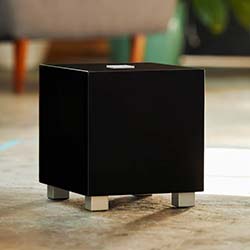 A sleek black cube speaker on a light-colored floor, with a blurred modern living room background featuring green plant leaves and wooden furniture legs.