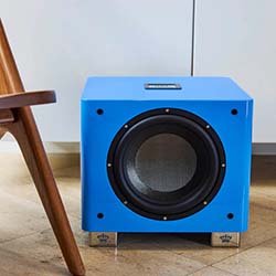 A bright blue subwoofer sits on a wooden floor next to a modern wooden chair, conveying a stylish, contemporary, and vibrant home audio setup.