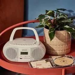 A vintage white boombox and potted plant sit on a round red table. Nearby are open CDs with colorful designs. The scene feels nostalgic and cozy.