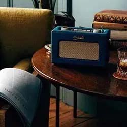 A cozy room with a vintage blue radio on a wooden table, beside a chair with an open magazine and a glass of whiskey, conveying a nostalgic vibe.