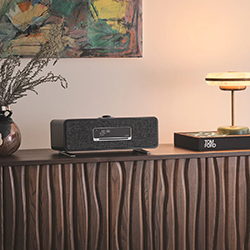 A wooden console with a wavy texture holds a vase with dried branches, a sleek speaker, and a modern lamp. Abstract art hangs on a warmly lit wall.
