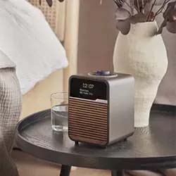 A sleek, modern alarm clock with a digital display reading "12:00" sits on a round table next to a glass of water and a ceramic vase with dried leaves.