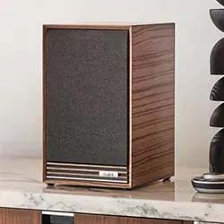 Vintage-style wooden speaker with a dark fabric front, set on a marble surface. Decorative black sculpture partially visible to the right. Elegant and retro.