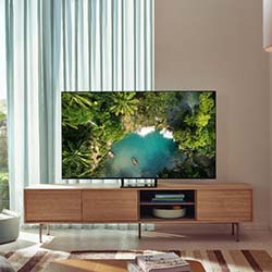 A flat-screen TV on a wooden stand displays a lush overhead view of a tropical paradise. Soft natural light filters through sheer curtains, creating a serene atmosphere.