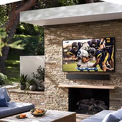 Outdoor patio with a stone fireplace featuring a large TV displaying a football game. Cozy seating area with cushions and a wooden table in the foreground.