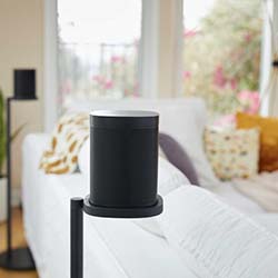 A sleek black speaker on a stand in a cozy living room with a white sofa, patterned and mustard pillows, large windows, and soft, natural lighting.
