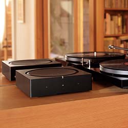 Three modern black audio devices on a wooden table, two with circular tops and a turntable with a record, set in a warm, cozy room with bookshelves.