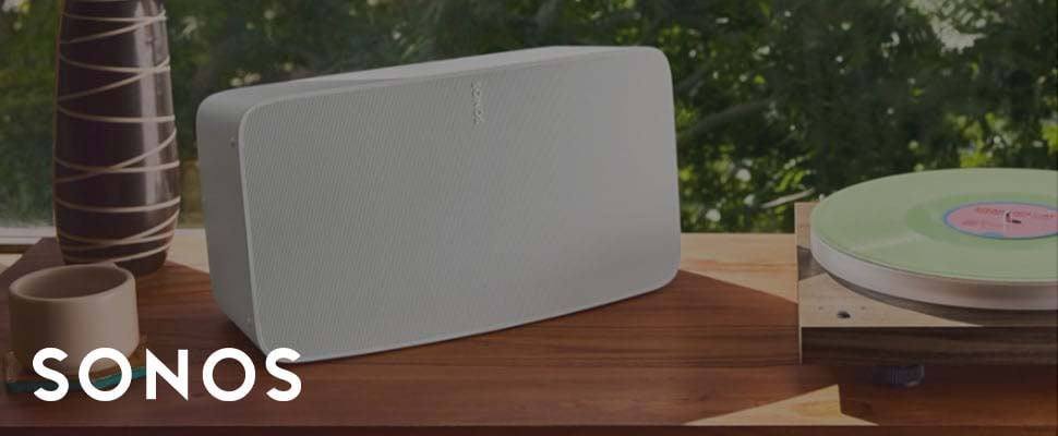 A white Sonos speaker is on a wooden table, next to a vase and a mug. A record player with a green vinyl is also present. The setting is calm and sunny.