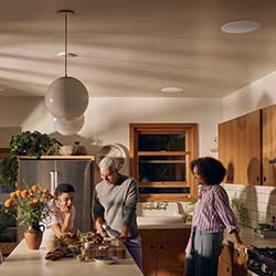 Three people gather in a cozy kitchen with warm lighting, preparing food together. There are wooden cabinets, a potted plant, and a sense of camaraderie.