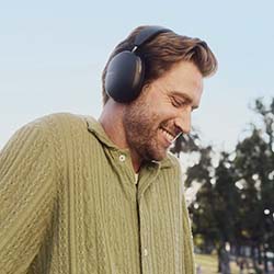 A smiling man in a green sweater enjoys music through headphones outdoors, with trees and a clear blue sky in the background, creating a relaxed, joyful scene.