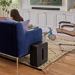 A cozy living room scene with a blue couch and a woman sitting, facing away. A sleek black subwoofer is nearby. A tricycle and elegant furniture adorn the space.
