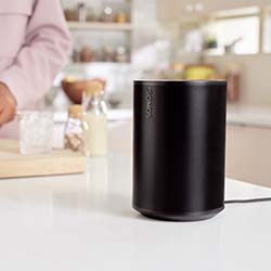 A black cylindrical smart speaker sits on a kitchen counter. In the background, a person is preparing food, creating a cozy, modern kitchen ambiance.