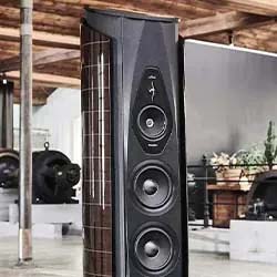A sleek black tower speaker with multiple drivers stands in a modern industrial space, featuring wooden beams and metal machinery, creating a sophisticated ambiance.