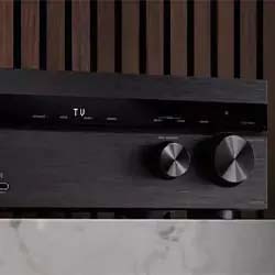 Close-up of a sleek, black audio receiver with two large knobs on the right and a digital display showing "TU." Set against a modern, wood-panel background.
