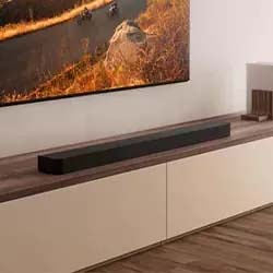 A sleek black soundbar rests on a wooden media console under a wall-mounted TV displaying a vibrant autumn landscape, creating a modern, cozy ambiance.
