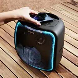 A hand places a smartphone on a black Sony wireless speaker with a glowing blue LED frame, resting on a wooden surface. The scene conveys a modern, tech-savvy ambiance.