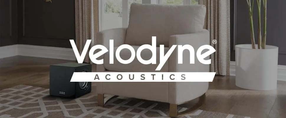 A cozy living room with a beige armchair on a patterned rug, next to a black subwoofer. The "Velodyne Acoustics" logo is prominently overlaid on the image.
