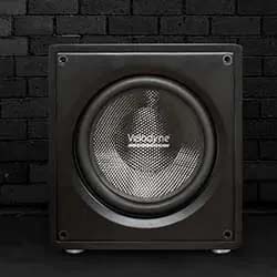 A black subwoofer with a prominent speaker grille, set against a dark brick wall. The scene conveys a sleek, modern, and powerful audio presence.
