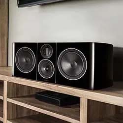 A black, sleek center speaker with three large drivers and a tweeter is displayed on a wooden shelf, suggesting a modern home audio setup.