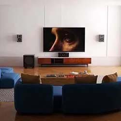 Spacious living room with a large TV displaying a close-up of a face. Blue sofas, wooden media unit, surround sound speakers, cozy and modern ambiance.