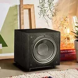 A black subwoofer sits on a cozy carpeted floor surrounded by books, a plant, and glowing lamp, creating a warm and inviting atmosphere.
