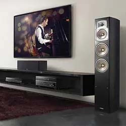 Living room with a wall-mounted TV displaying a pianist. A tall speaker stands nearby, conveying a sense of elegance and rich sound quality.