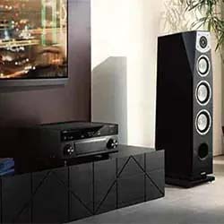 A modern living room with a large black speaker tower, a TV on the wall, and a stereo receiver on a sleek black cabinet. The scene conveys a high-tech, elegant vibe.