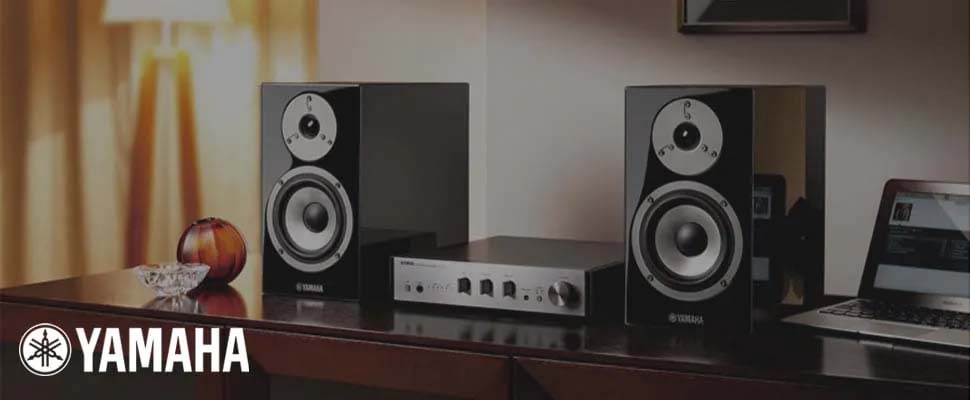Two sleek Yamaha speakers flank a central audio amplifier on a wooden table, creating a modern and elegant sound setup. A laptop and decorative items add warmth.