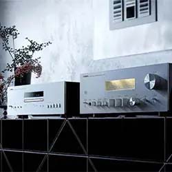 Vintage audio equipment on a black shelf, featuring a CD player and an amplifier with knobs. A small potted plant and a vented wall add a retro vibe.