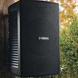 Outdoor speaker mounted on a wooden post, surrounded by greenery. The black speaker has the Yamaha logo, set against a blurred natural background.