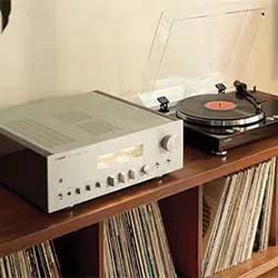 A vintage turntable with a vinyl record and an audio receiver sit atop a wooden shelf filled with records, evoking a nostalgic, retro vibe.