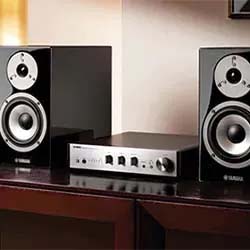 A sleek audio setup featuring two black Yamaha speakers flanking a modern amplifier on a wooden surface, set against a softly lit beige wall.