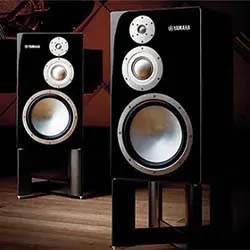 Two sleek Yamaha speakers in a dimly lit room, displaying a modern design with large woofers and tweeters, convey a sophisticated audio setup.