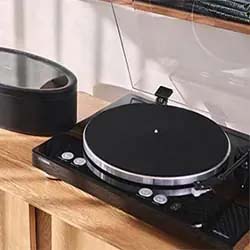 A sleek black turntable sits on a wooden shelf next to a round black speaker. Sunlight casts soft shadows, creating a calm, nostalgic ambiance.
