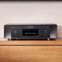 A sleek black audio receiver with front display and controls sits on a wooden shelf. The digital screen shows text, creating a modern, sophisticated ambiance.