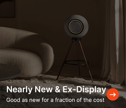 A modern speaker on a tripod stands beside a curved beige sofa in a softly lit room. Text reads: "Nearly New & Ex-Display. Good as new for a fraction of the cost."