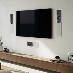 Wall-mounted flat-screen TV with three sleek speakers around it on a minimalist white wall. Below, a wooden shelf holds decor for an elegant setup.