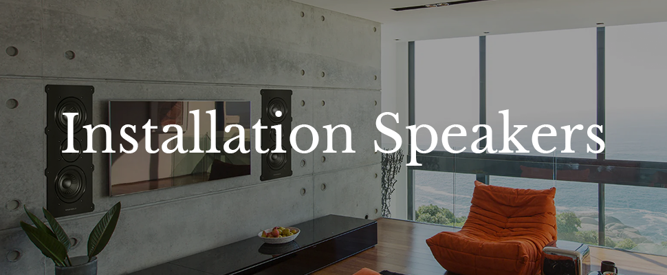 Modern living room with large windows and ocean view. Wall-mounted speakers flank a flat-screen TV on a concrete wall. Text reads "Installation Speakers."