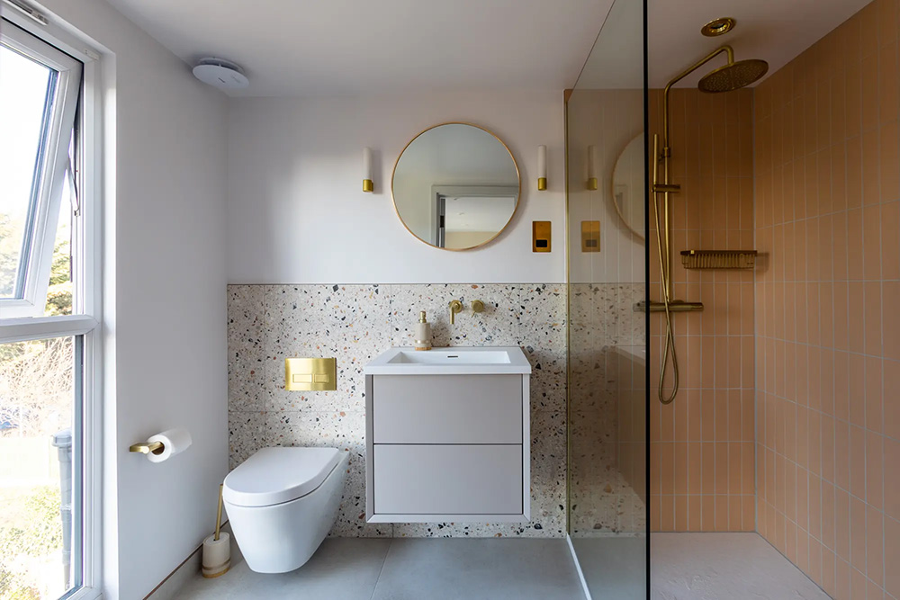 Modern bathroom with terrazzo wall, white fixtures, gold accents, and a glass shower. Round mirror and light fixtures enhance the clean, elegant look.