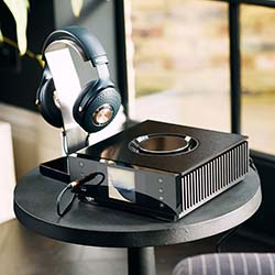 High-end headphones rest on a sleek, black audio amplifier placed on a round table by a sunlit window. The setting conveys a modern, relaxing atmosphere.