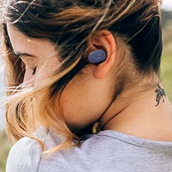 A woman with tied hair, wearing a gray shirt, listens to music with wireless earbuds. Her head is turned, showing a neck tattoo and conveying a serene mood.